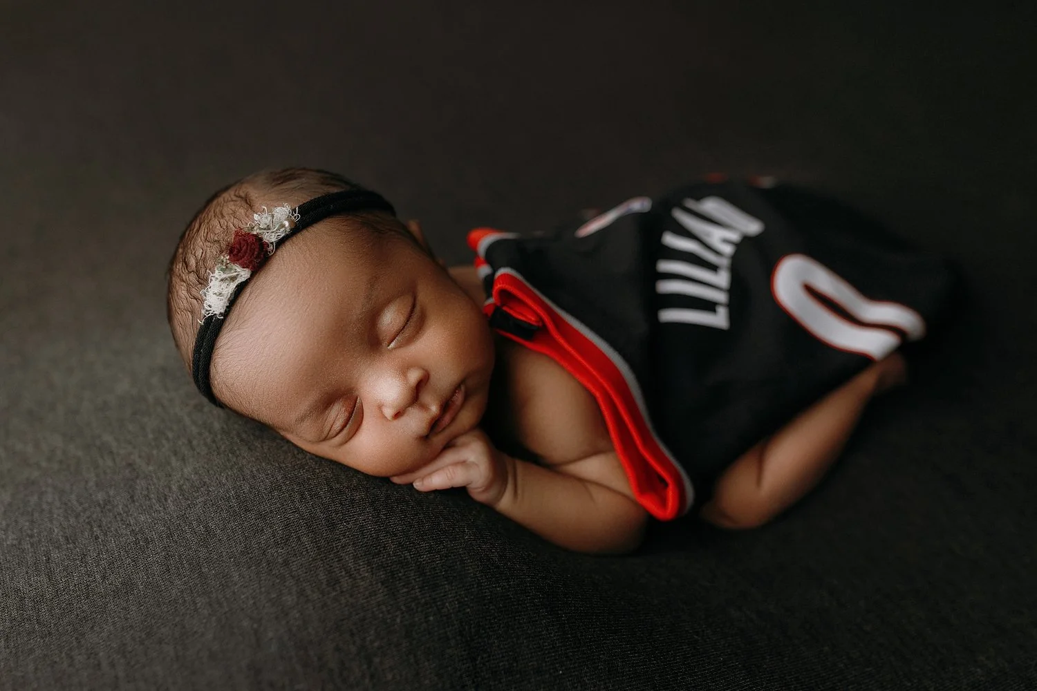 A professional studio newborn portrait by SunnyMelon Photography featuring Damian Lillard's twin daughter. The sleeping infant is posed delicately on her stomach against a dark charcoal textured backdrop. She is draped in a black, red, and white Port