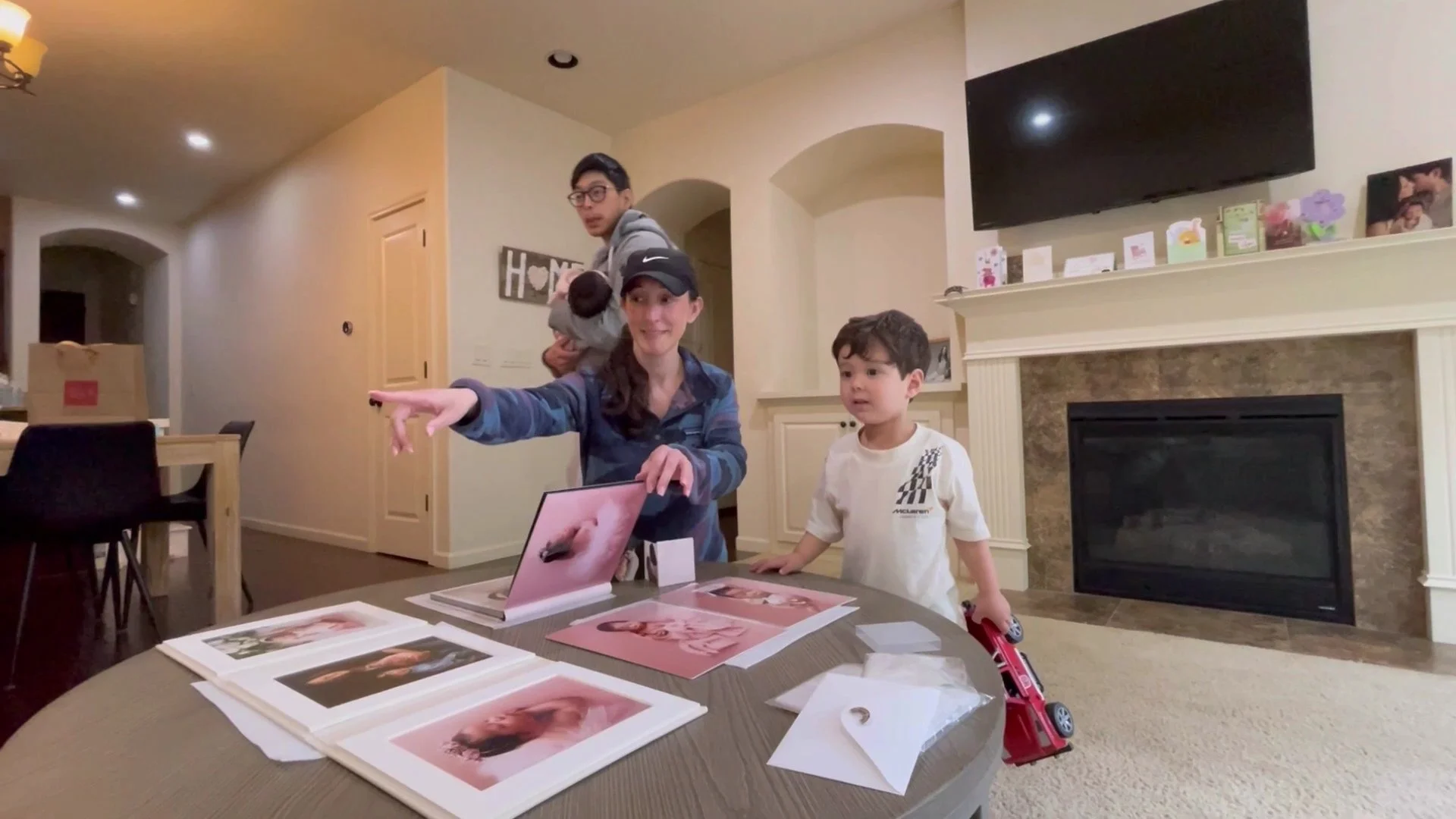 A family of four, including a young boy and a father holding an infant, gathered around a coffee table looking at professional newborn photo prints in a living room.