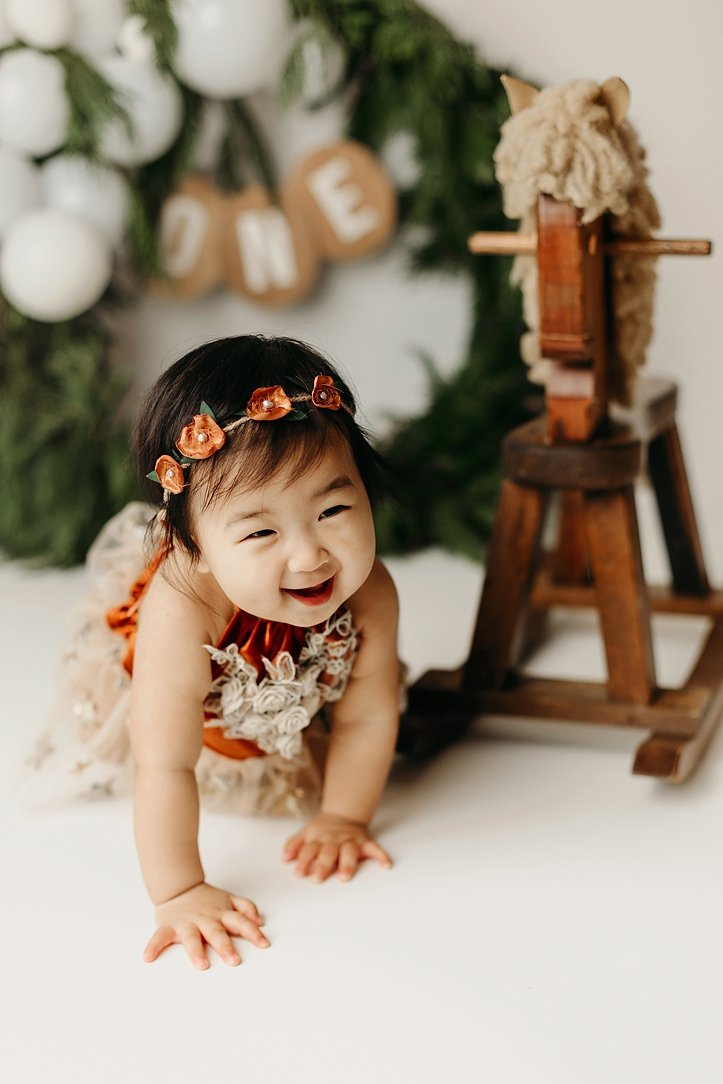 This heartwarming milestone portrait features a happy baby girl celebrating her first birthday with a nature-inspired studio theme. She is captured mid-crawl, wearing a delicate orange floral headband and a coordinated dress with lace detailing. The 