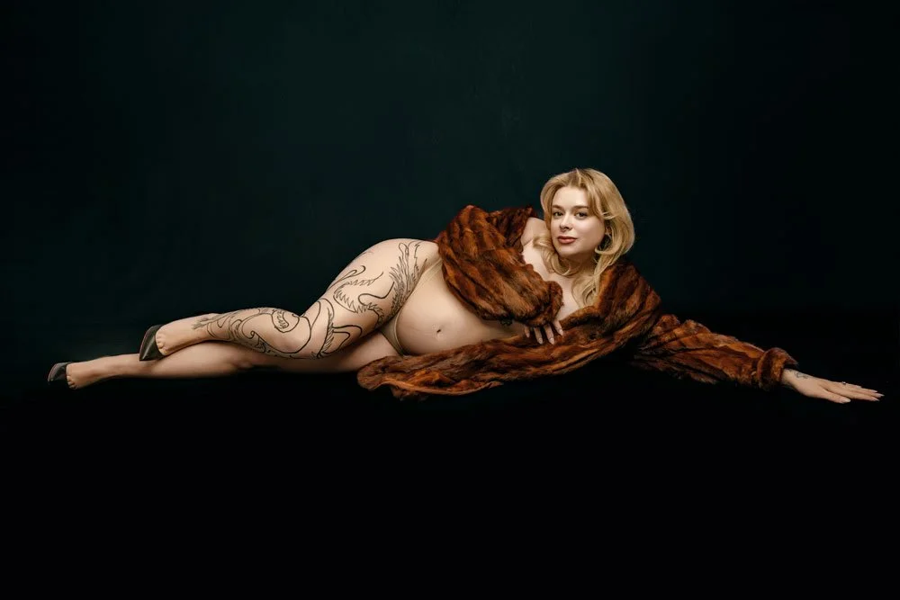 A luxury maternity boudoir portrait of a pregnant woman reclining against a black backdrop in a West Linn studio. Draped in a rich fur coat with an artistic leg tattoo, she elegantly showcases her baby bump.