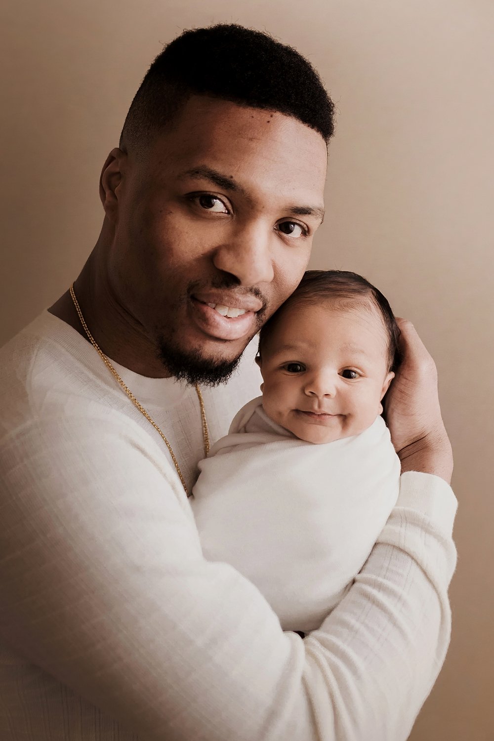 A warm and intimate studio newborn portrait by SunnyMelon Photography featuring NBA star Damian Lillard holding his son, Dame Jr. The baby is snugly wrapped in a white swaddle, smiling softly at the camera while Damian cradles his head and looks on w
