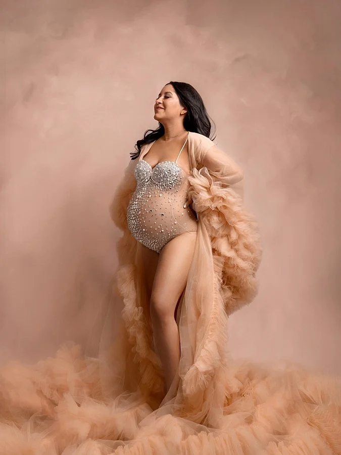 Sunnymelon Photography ultra-glamorous and joyful maternity portrait. An expectant mother is posing in a sparkling, rhinestone-embellished bodysuit layered with a dramatic, ruffled tulle robe in a peach hue, smiling with her eyes closed as she celebr
