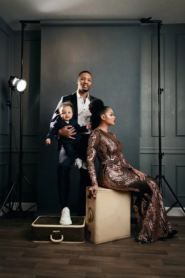 A professional studio portrait of Damian Lillard holding his young son while standing behind his partner, Kay'La, who is seated on a vintage cream suitcase. They are in formal attire against a charcoal gray backdrop with studio lighting visible.