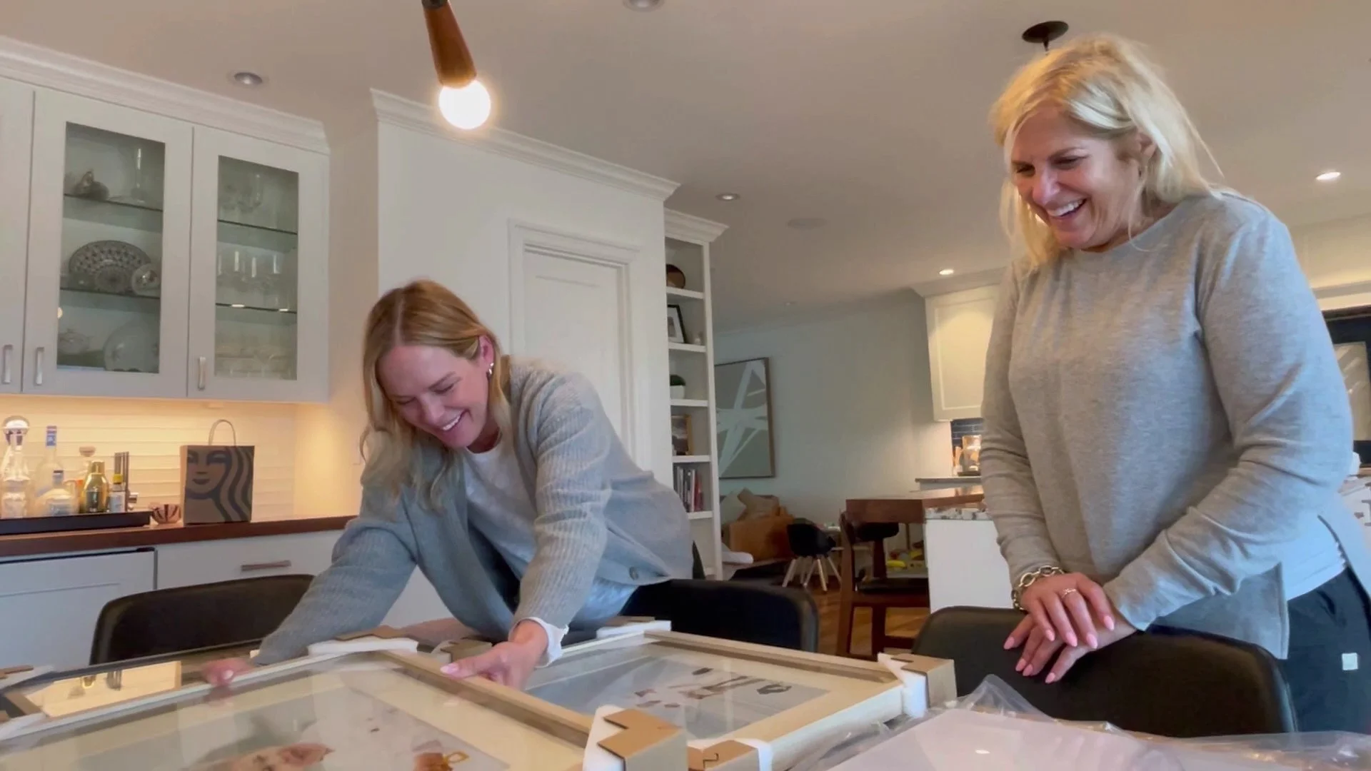 Two smiling women in a modern kitchen unboxing large, professionally framed photo prints with protective corner packaging.