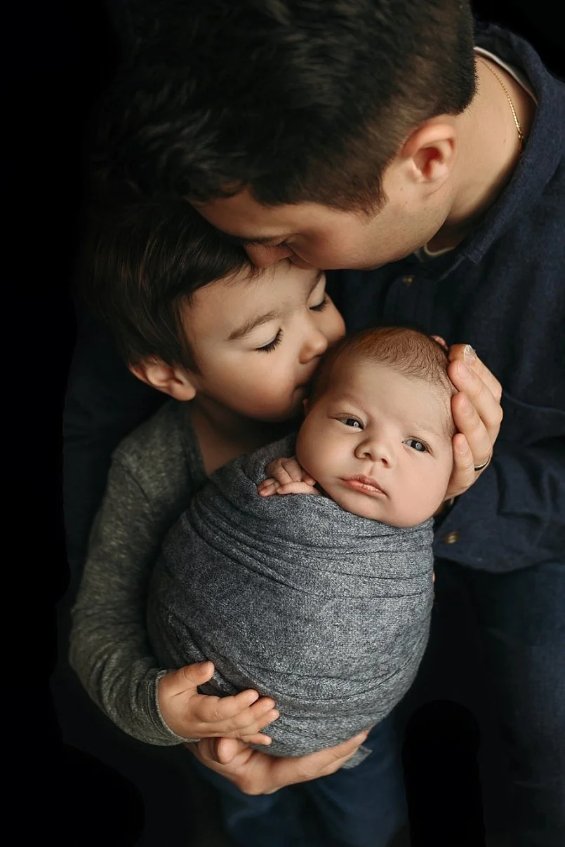 A soulful and moody studio newborn portrait by SunnyMelon Photography featuring an older brother gently kissing his swaddled baby brother's head. The newborn is snugly wrapped in a textured charcoal gray swaddle. Both are being held by their father, 