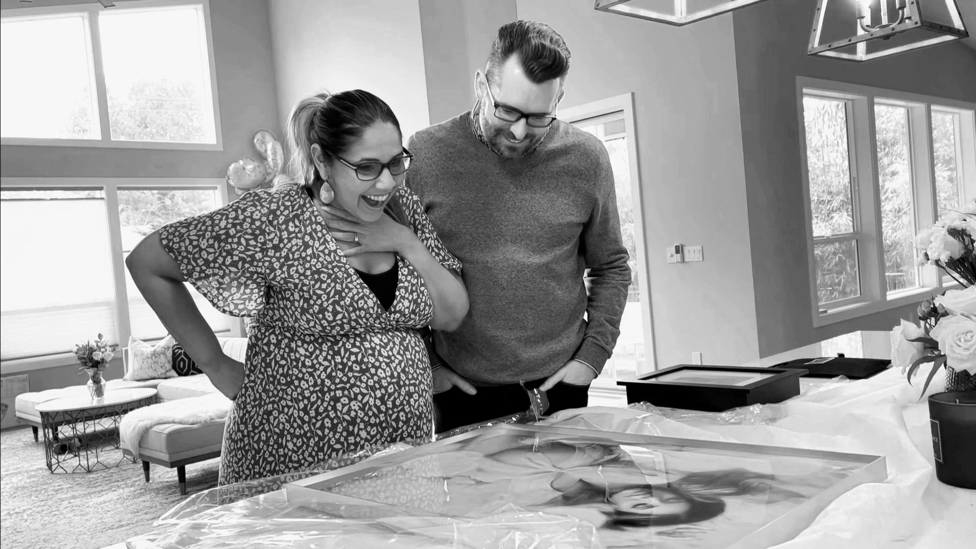 A black and white photo of an expectant mother and father reacting with joy and excitement while unboxing a large, framed fine-art portrait on their kitchen counter.