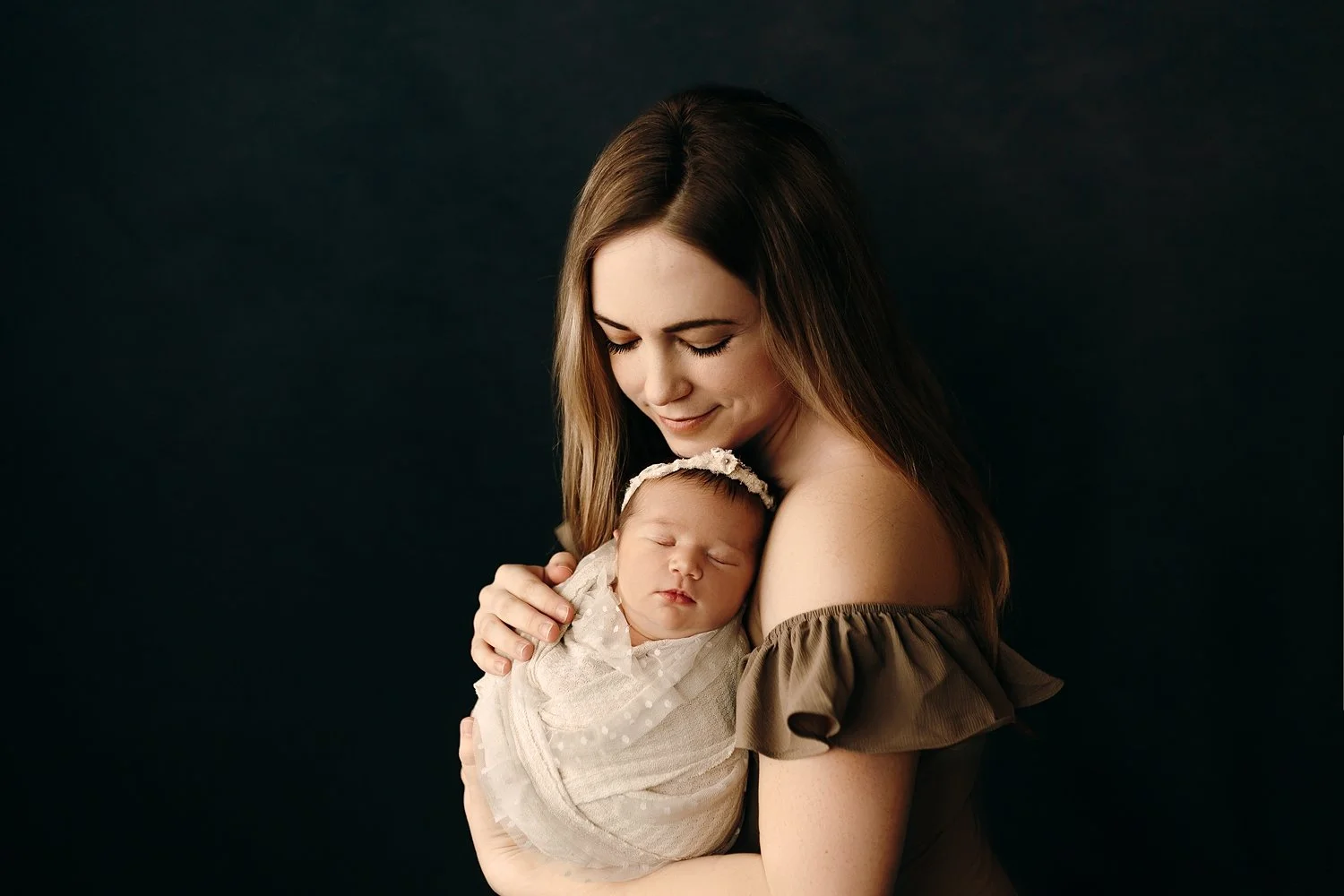 A heartwarming newborn photograph featuring a woman in an off-the-shoulder olive green top gently holding an infant. The baby is peacefully asleep, wrapped in a white lace swaddle with a delicate floral headband. The mother’s eyes are closed as she r