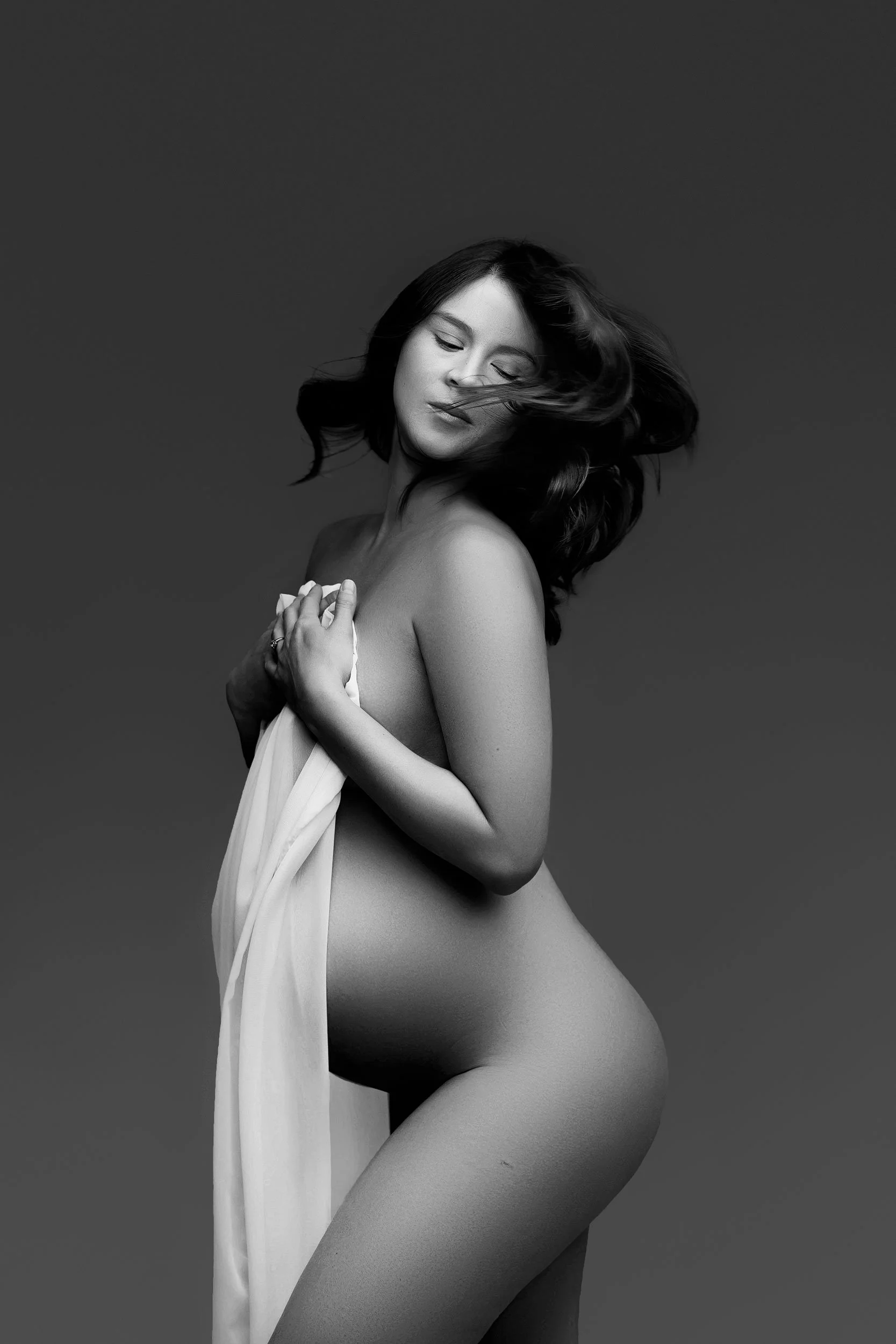 Sunnymelon Photography exquisite black and white fine art maternity portrait. The expectant mother is partially draped in a sheer white fabric, with her hair dramatically windblown, eyes closed in a serene expression, highlighting her pregnant belly.