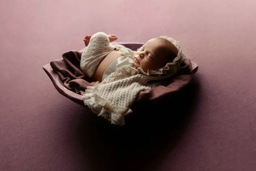 A sleeping baby lying on a cushioned purple cradle, dressed in a white knitted outfit with a bonnet, against a soft pink background.