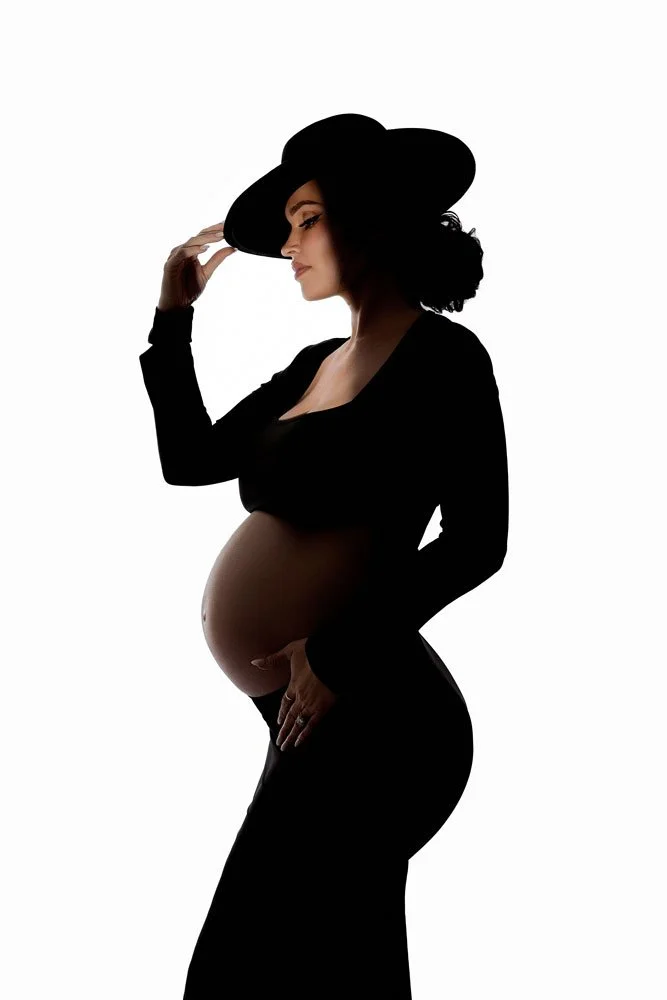 A striking fine-art maternity silhouette portrait of a pregnant woman in profile against a bright white backdrop in West Linn, Oregon. Styled in a black crop top, skirt, and fedora, she tips her hat while cradling her baby bump.