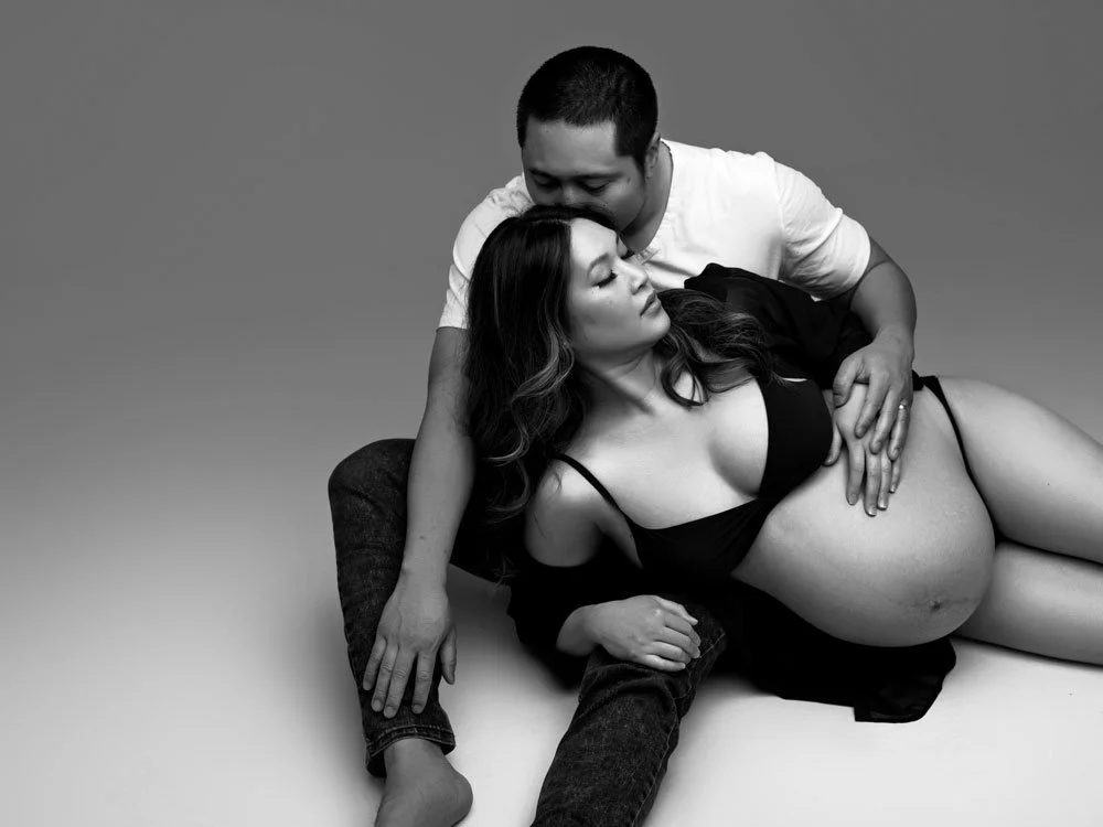 Sunnymelon Photography intimate black and white couples maternity portrait. The expectant mother is lying on her back in a bra, cradling her pregnant belly, while her partner gently kisses her head. They are seated closely on the floor.