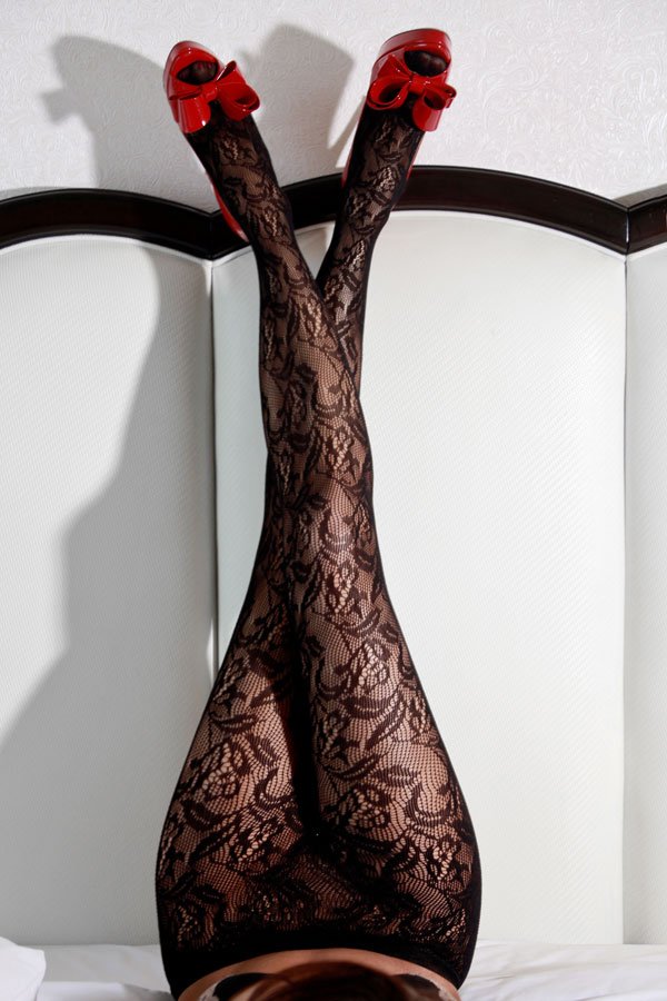 A woman's legs in intricate floral lace tights and bright red high-heeled shoes with bows, resting vertically against a white tufted headboard.