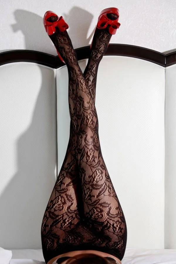 A woman's legs in intricate floral lace tights and bright red high-heeled shoes with bows, resting vertically against a white tufted headboard.