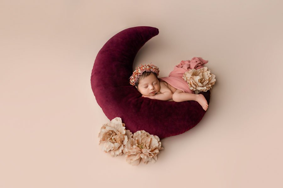 A whimsical newborn photography setup featuring an infant peacefully sleeping on her stomach atop a deep burgundy crescent moon-shaped cushion. The baby wears a dainty crown of small pink and orange flowers and is partially draped in a dusty rose fab