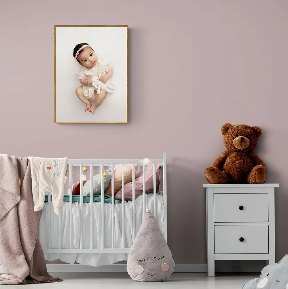 Gold framed vertical fine-art baby portrait mockup hanging on a dusty rose nursery wall above a white crib.