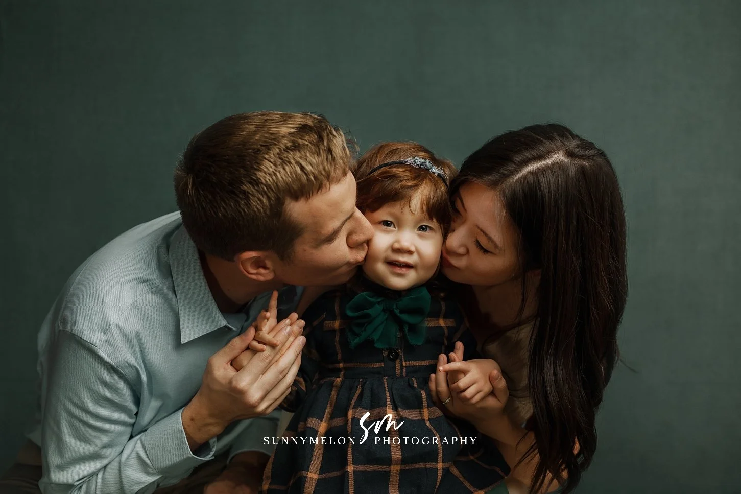 A professional studio portrait of a mother and father kissing their toddler daughter on the cheeks against a textured deep green backdrop, showcasing a tender family moment.