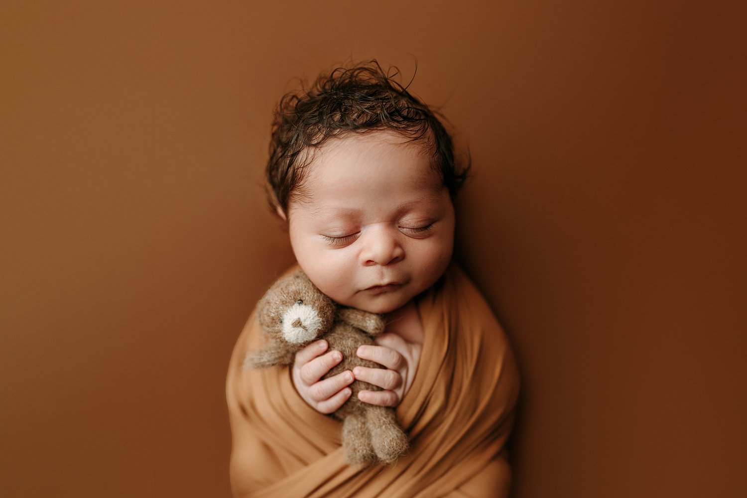 A serene, monochromatic newborn photograph featuring an infant peacefully sleeping. The baby has soft, curly dark hair and is tightly wrapped in a caramel-colored fabric. Cradled in the baby's small hands is a miniature, brown fuzzy teddy bear. The e