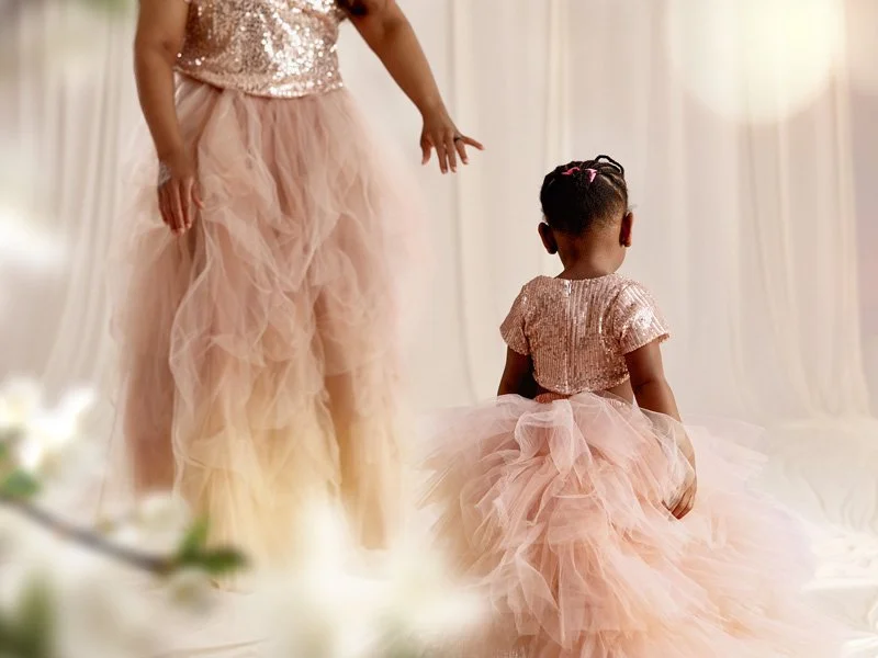 A young girl viewed from behind, wearing a rose gold sequin top and a voluminous blush pink tulle skirt, standing in a bright, ethereal studio with her mother's skirt visible in the background.
