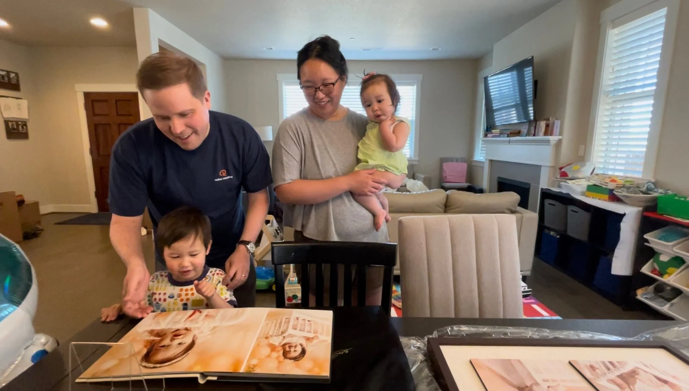 A family of four inside their living room gathered around a dining table, smiling as they look through a large, high-end flush-mount photo album alongside packaged framed wall art prints.