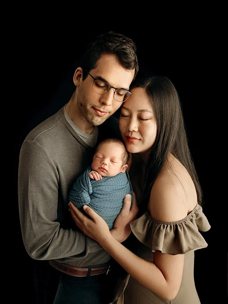An intimate family photograph featuring a man with glasses in a grey sweater and a woman with long dark hair in an off-the-shoulder olive top. They are positioned close together with their foreheads nearly touching, looking down at their infant who i