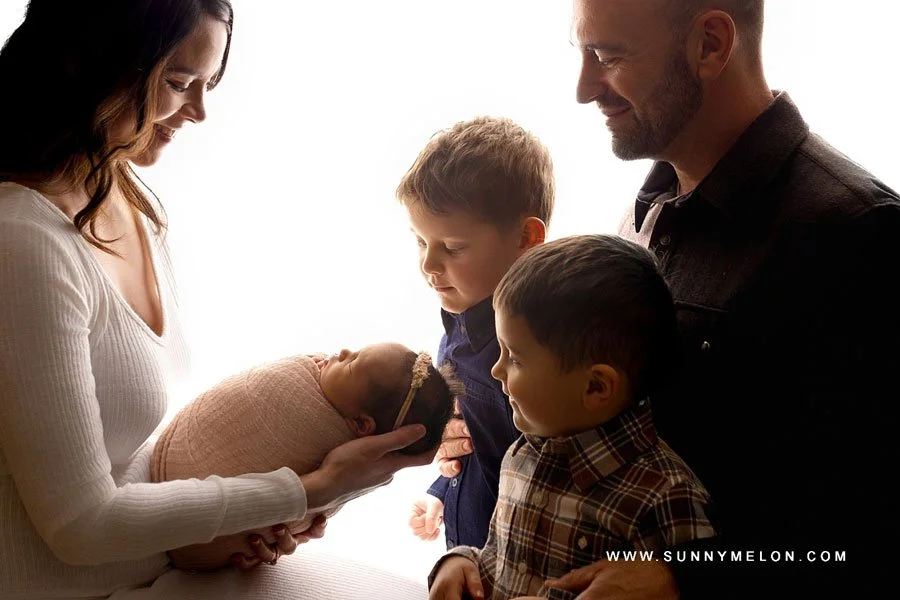 A back-lit studio family portrait where a mother in white holds a swaddled newborn baby, while the father and two young sons look on lovingly against a bright white background.
