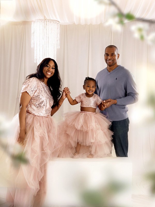 A happy family of three, with a woman, a man, and a young girl, standing together indoors against a white backdrop. The young girl is in the middle wearing a pink tulle dress, holding hands with both adults who are smiling.