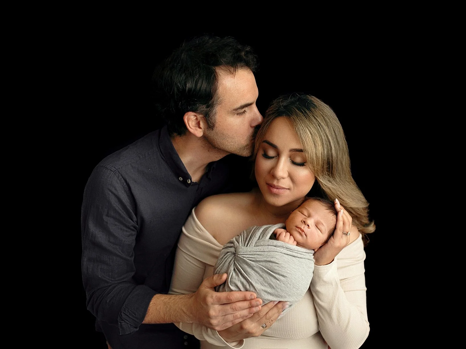A classic fine art studio newborn portrait featuring a mother and father holding their sleeping baby against a solid black background. The father is leaning in to kiss the mother’s forehead while she cradles the infant, who is snugly wrapped in a sof