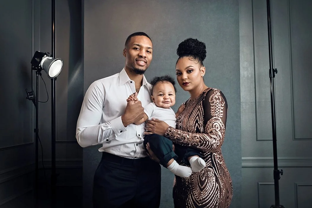 This elegant family portrait by SunnyMelon Photography features NBA star Damian Lillard in a white dress shirt, Kayla Lillard in a shimmering gold sequin gown, and their son, Dame Jr.. The image is captured in a professional studio setting with visib