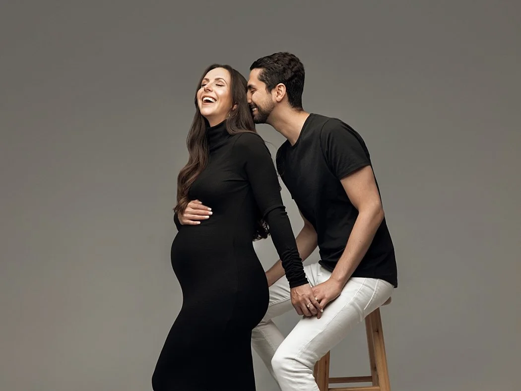 A candid studio maternity portrait of a laughing couple against a grey backdrop. The expectant mother stands in a fitted black dress, while her partner sits on a wooden stool in a black t-shirt and white pants.