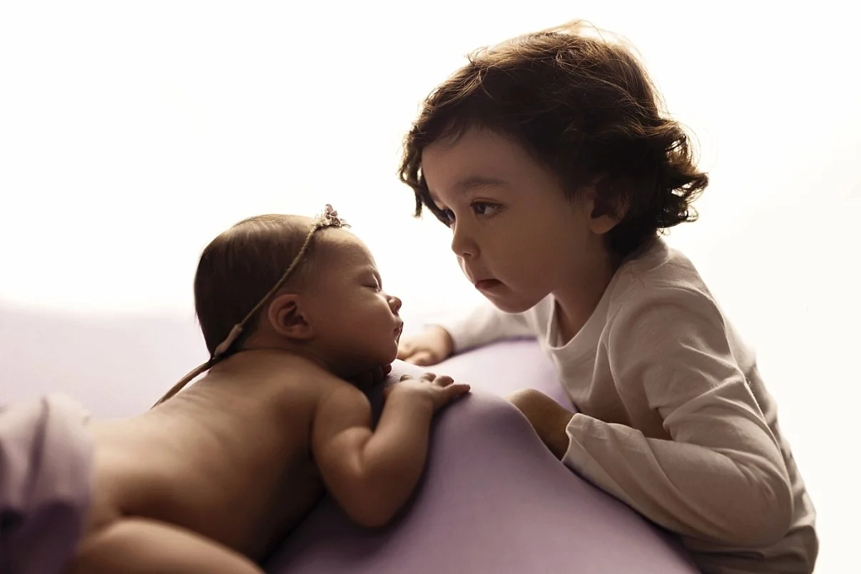 A tender sibling moment captured in a high-key photography style. An older toddler boy in a white long-sleeve shirt leans in to look closely at his newborn sister. The baby is peacefully asleep on her stomach, resting on a lavender or light purple be