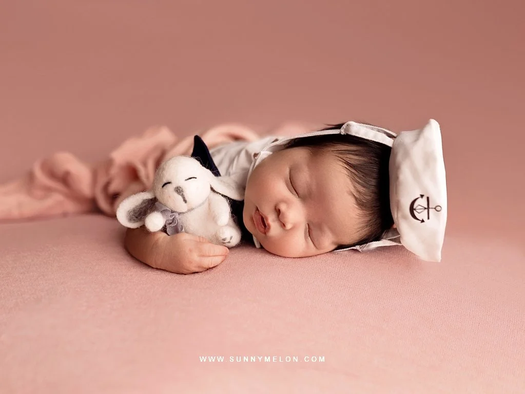 A side-profile of a sleeping newborn baby girl on her side, wearing a small white sailor hat with an anchor, holding a plush white bunny, set against a monochromatic pink studio background.