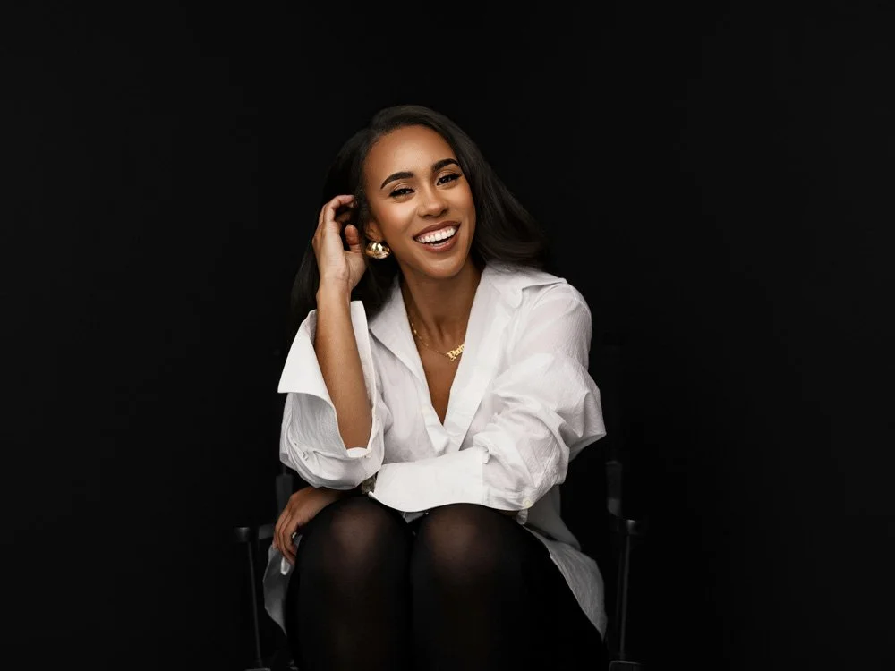 A professional woman with long dark hair, wearing a white dress shirt and black leggings, smiles while seated in a black director's chair against a solid black studio background.