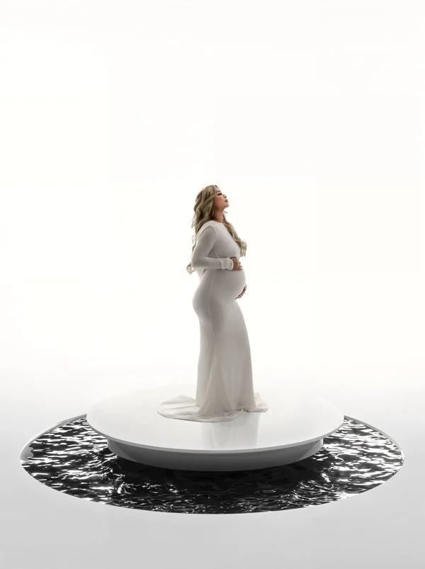 A pregnant woman with long blonde hair wearing a fitted white long-sleeved gown, standing on a white circular platform surrounded by a dark reflection pool against a bright white background.