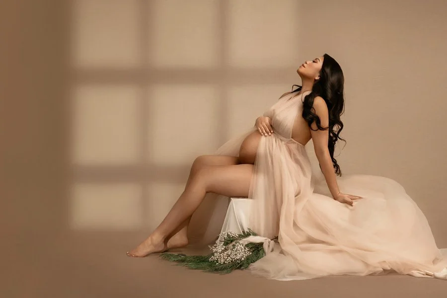A high-end studio maternity portrait of a pregnant woman with long dark hair, wearing a flowing sheer peach tulle gown, reclining on a white pedestal against a warm-toned background with window light shadows.