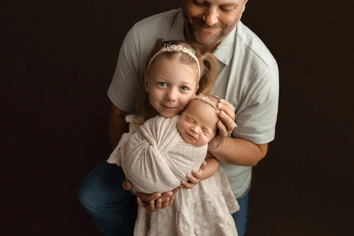 A fine-art studio portrait of a father and young daughter cradling a swaddled, sleeping newborn against a dark brown backdrop in West Linn, Oregon. The warm family moment is captured under soft, high-end lighting.
