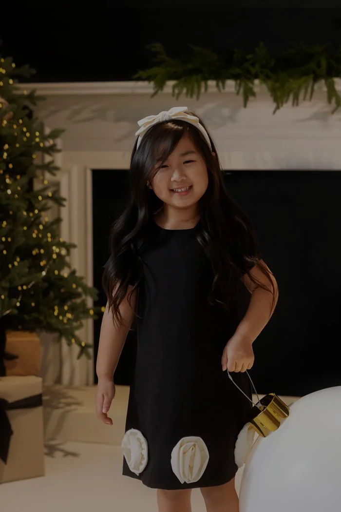 A young girl smiling during a holiday mini session in Oregon. She is wearing a black dress with white floral accents and a white headband, standing in a decorated studio featuring a lit Christmas tree and a minimalist fireplace mantle.