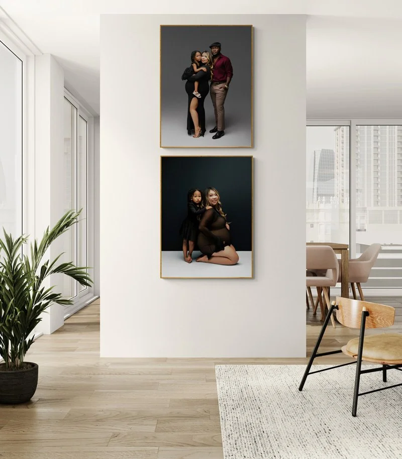 A bright, modern living room featuring two large framed vertical portraits on a white wall: a family maternity pose in the top frame and a mother-daughter maternity portrait in the bottom frame.