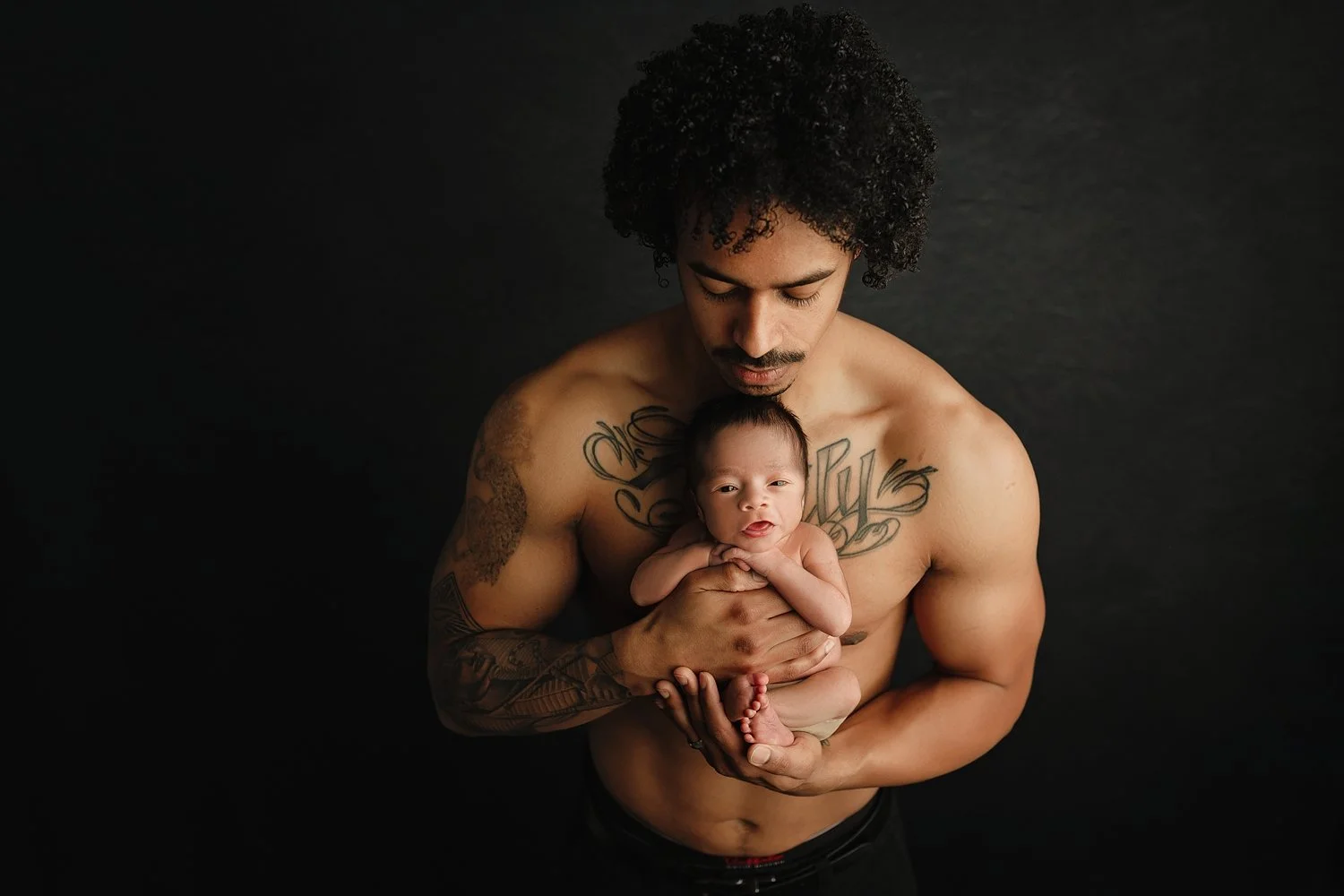 An intimate, low-key newborn photograph featuring a father with dark curly hair looking down at the infant he holds in his hands. The newborn is awake, looking toward the camera while nestled in its father's palms. The father's large chest tattoo is 