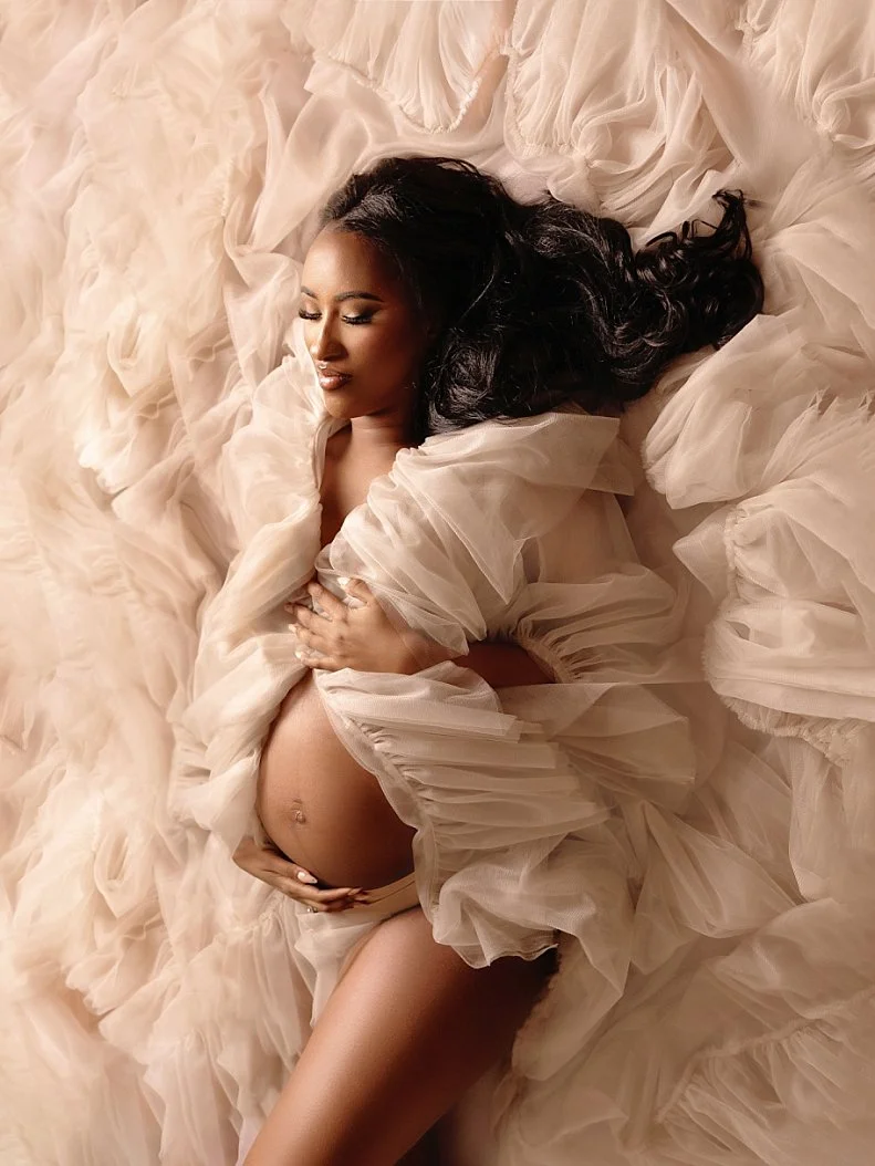 Beautiful maternity photoshoot in West Linn, Oregon, with a pregnant woman lying down in a soft, ruffled white gown, gently holding her baby bump.