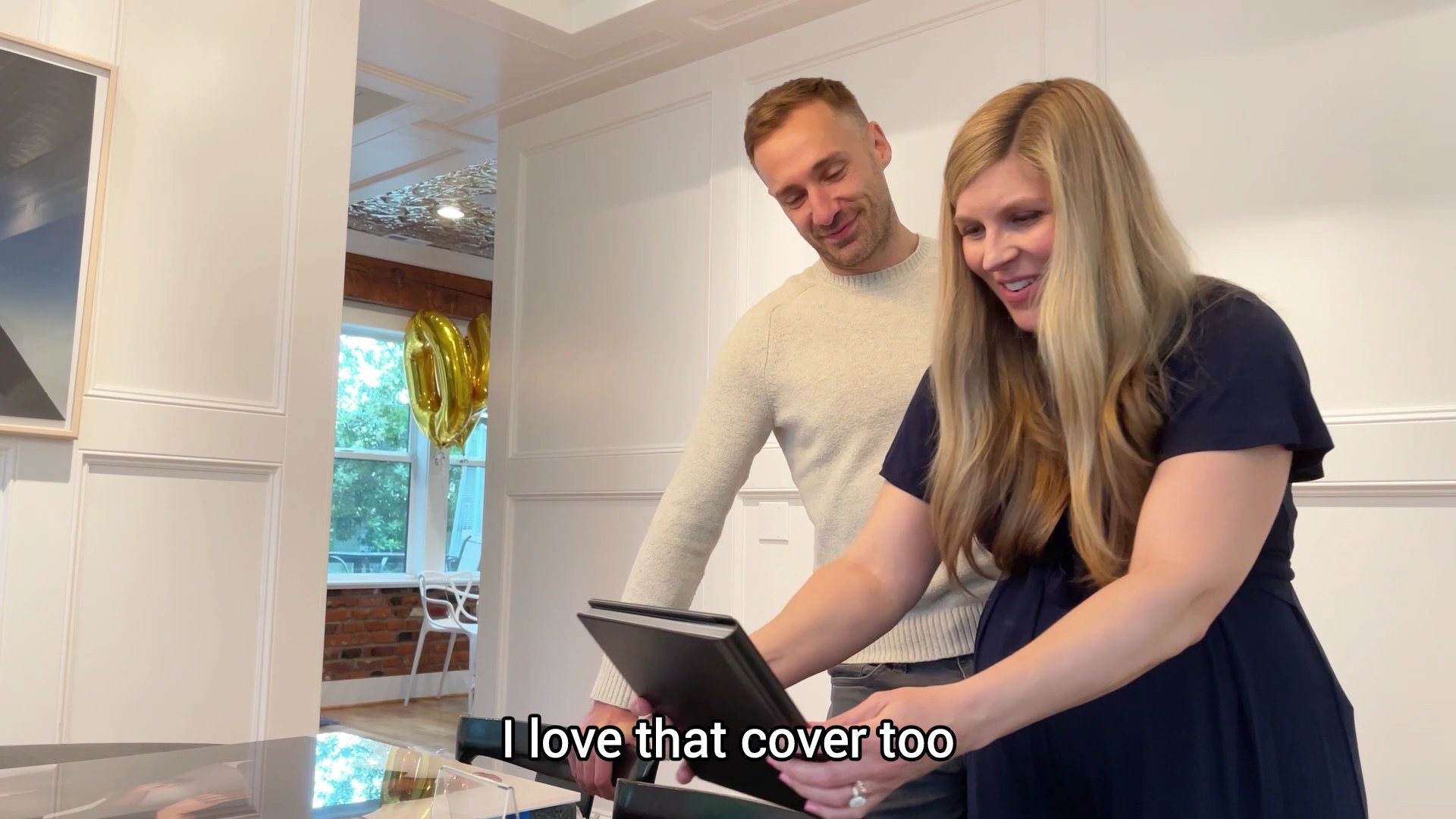 A man and a woman are shown in a modern, bright interior, smiling as they examine the cover of a professional photography album. The woman holds the black, high-end photo book while the man looks on with a pleased expression. This image captures a "r