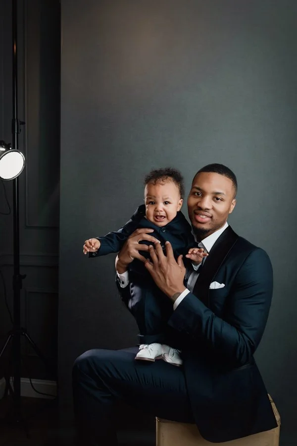 Professional NBA player Damian Lillard in a black tuxedo sitting and holding his young son, also in a matching suit, against a dark gray studio backdrop with professional lighting.