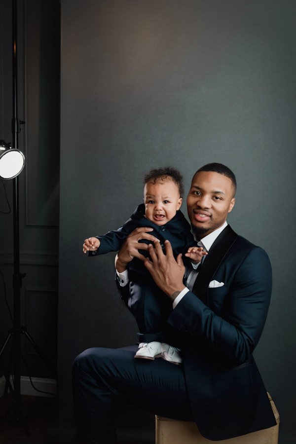 Professional NBA player Damian Lillard in a black tuxedo sitting and holding his young son, also in a matching suit, against a dark gray studio backdrop with professional lighting.