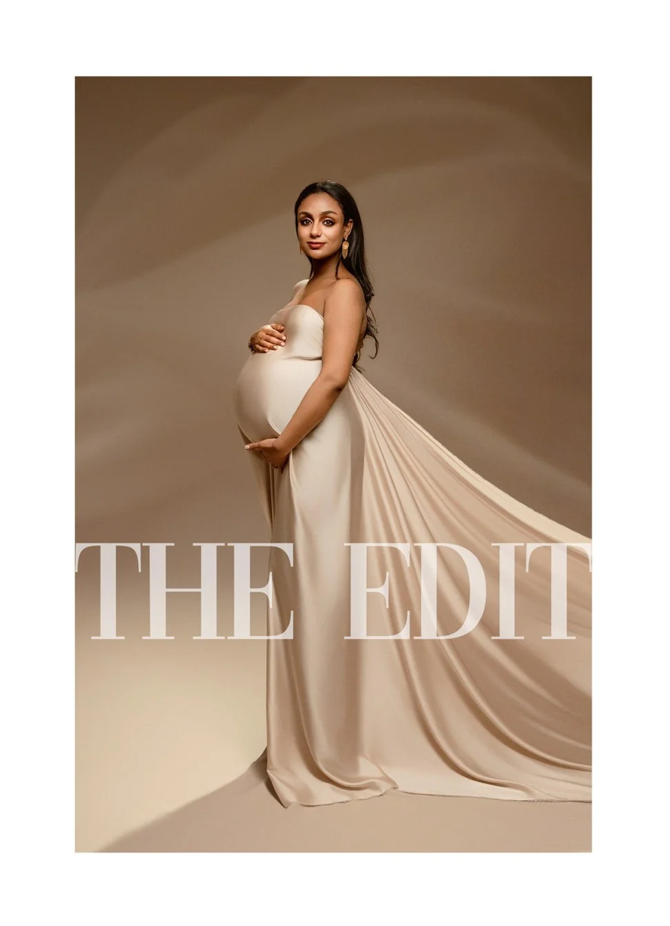 A professional studio maternity portrait of a pregnant woman wearing a flowing, strapless champagne satin gown with a long train, posing against a warm neutral backdrop.