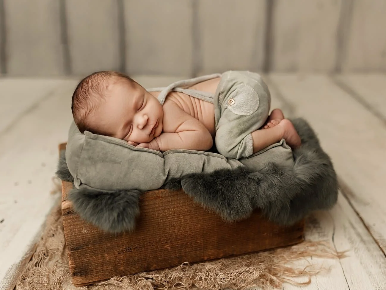 A newborn baby boy wearing sage green knit overalls sleeps peacefully in a bum-up pose. He rests on a matching green pillow and faux fur lining inside a rustic wooden crate, set against a light wood-plank backdrop.