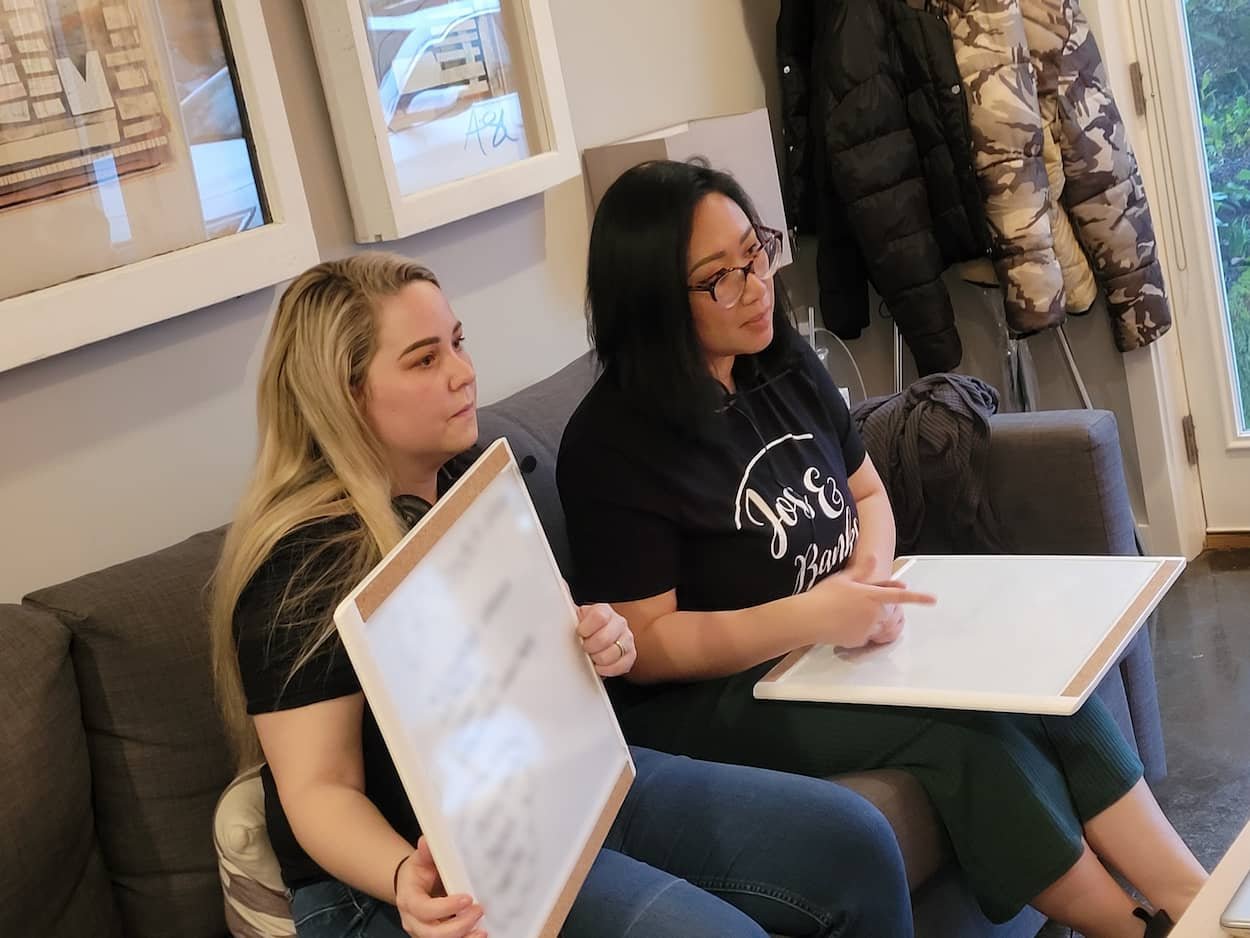 Josie Tan and Kristina Banks sitting on a grey couch, holding whiteboards while filming educational content for their photography mentor program.