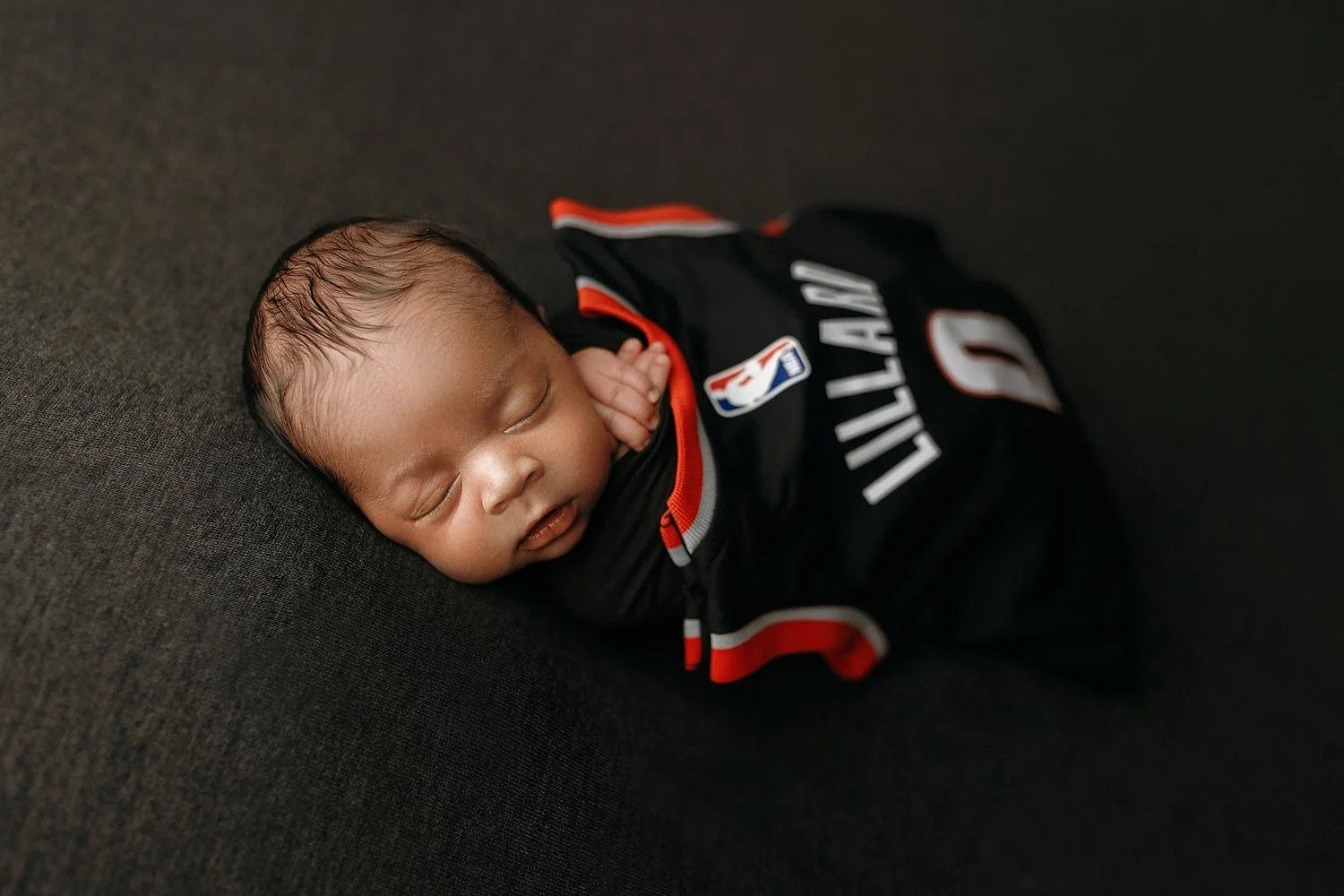 A professional studio newborn portrait by SunnyMelon Photography featuring Damian Lillard's twin son. The sleeping infant is posed delicately on a dark charcoal textured surface. He is draped in a black, red, and white Portland Trail Blazers jersey f