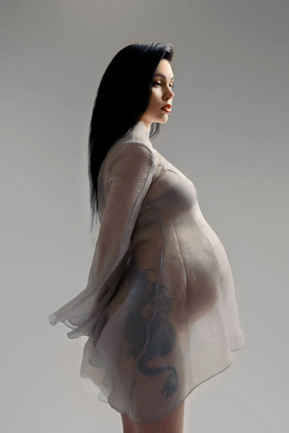 An elegant fine-art studio maternity portrait of a pregnant woman in profile against a grey backdrop in West Linn, Oregon. She wears a delicate, sheer grey fabric that drapes over her baby bump, highlighting her silhouette under soft studio lighting.