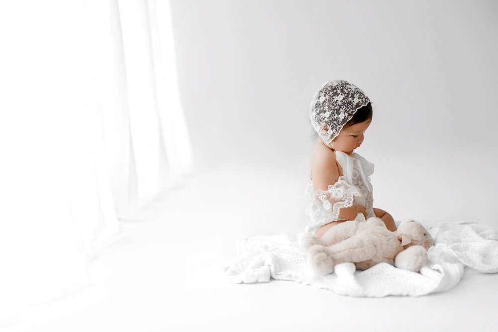 A young baby sitting on a white blanket in a bright, ethereal studio, wearing a delicate white lace bonnet and matching outfit while playing with a plush teddy bear.