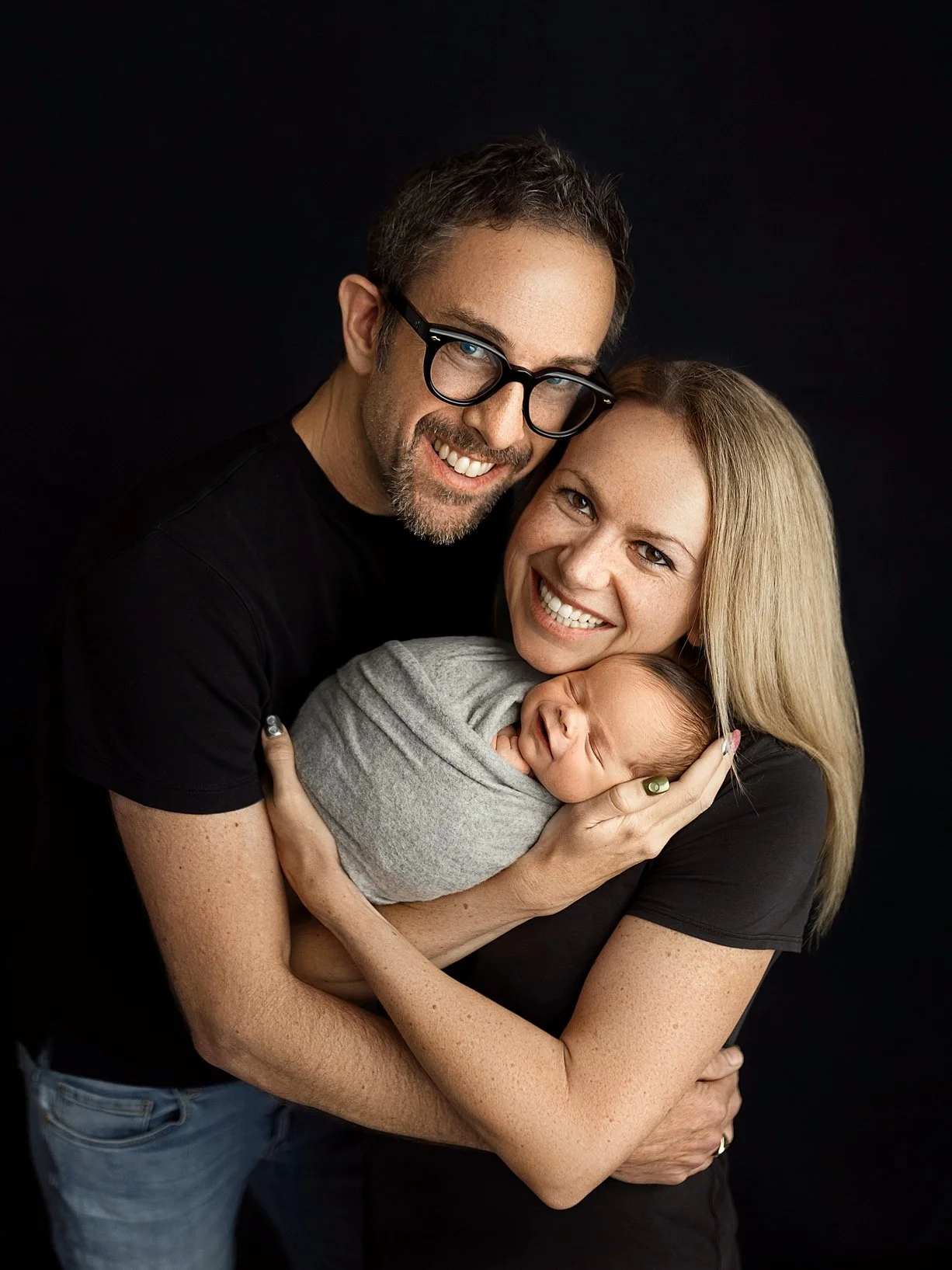 A heartwarming family photograph featuring a blonde woman and a man with glasses, both smiling radiantly at the camera. They are closely embracing their newborn infant, who is snugly swaddled in a soft grey wrap and appears to be smiling in its sleep