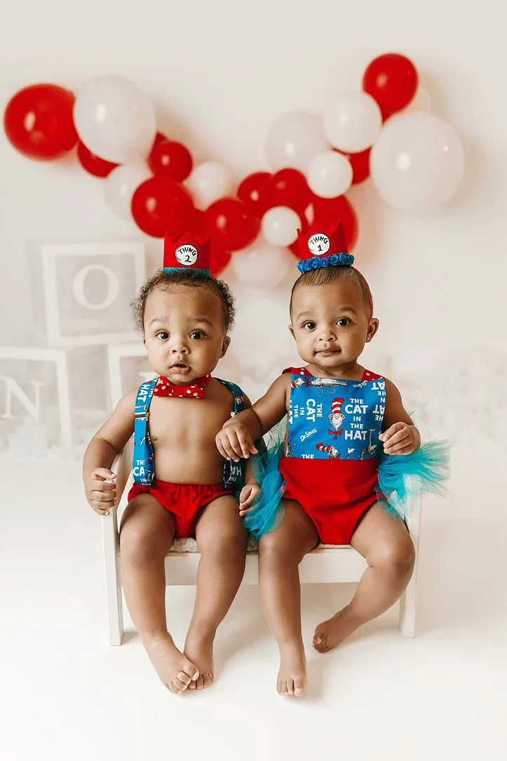 This adorable milestone portrait features twin babies celebrating their first birthday with a vibrant Dr. Seuss "Thing 1 and Thing 2" theme. The infants are posed on a miniature white bench, wearing coordinated red and blue outfits complete with them
